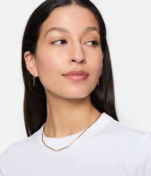 Goudkleurige geometrische oorbellen en een bijpassende ketting met cilindrische en ronde vormen. De sieraden contrasteren met het witte shirtstof.
