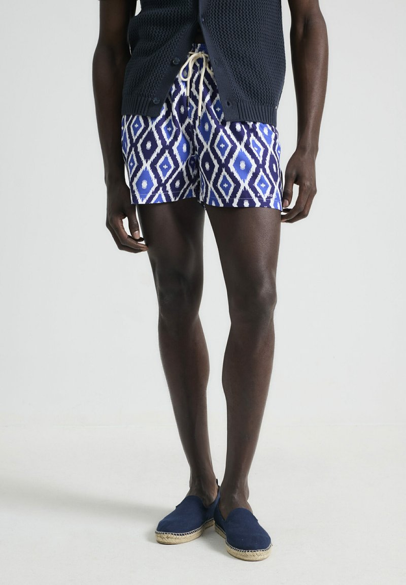 Bañador azul con estampado y formas de diamante en blanco, con cintura elástica y cordón ajustable, combinado con espadrilles marinos, sobre un fondo neutro.