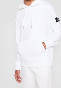 Vit bomullshuvtröja med framficka, försedd med dragsko och en svart märkesetikett på vänster ärm. Mjuk textur.