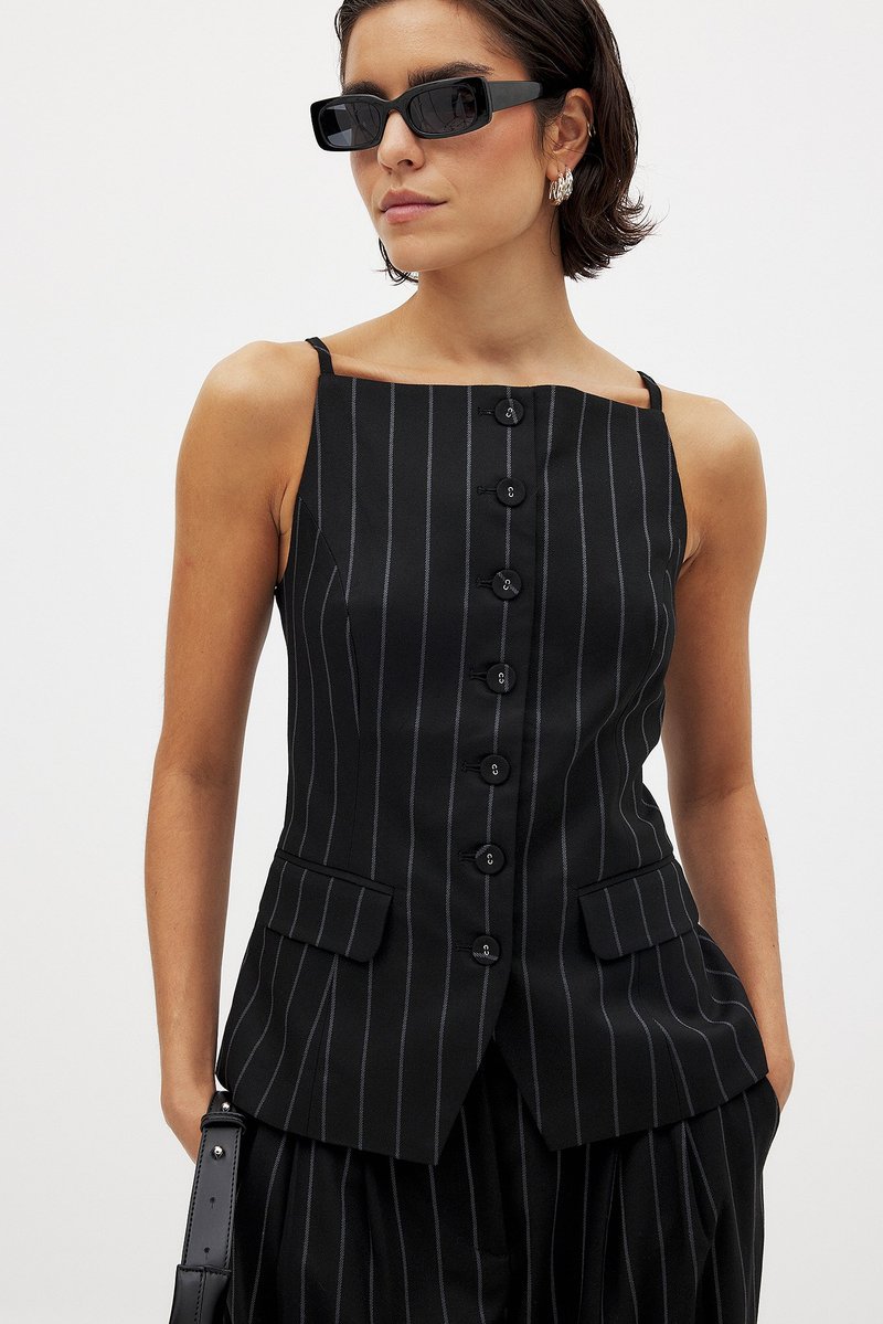 Ženska nosi črn prugast brezrokavnik s gumbi in kvadratnim izrezom, usklajene prugaste hlače, črne sončne očala in srebrne obroče.