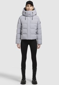 Hellgraue Steppjacke mit hohem Kragen, voluminösem Steppmuster, vorderen Knöpfen und Kordeln. Getragen über schwarzen Leggings und Stiefeln.