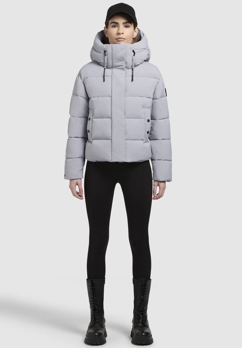 Hellgraue Steppjacke mit hohem Kragen, voluminösem Steppmuster, vorderen Knöpfen und Kordeln. Getragen über schwarzen Leggings und Stiefeln.
