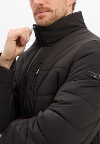 Czarna puchowa kurtka z wysokim kołnierzem, zamkiem z przodu i pikowaną teksturą. Posiada naszywkę z logo na rękawie oraz srebrne suwaki.