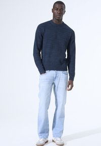 Marineblauwe gebreide trui met een gestructureerd patroon, ronde halslijn en geribde manchetten, gecombineerd met lichtblauwe jeans en grijze sneakers.