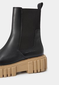 Bottine en cuir noir avec panneau latéral élastique et semelle beige épaisse à crampons, avec une languette à l'arrière.