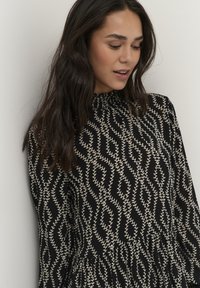 Zwarte jurk met een gestructureerd patroon van crème bladeren, een geplooide halslijn en lange mouwen. Een uitlopende zoom geeft het ontwerp extra beweging.