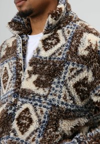 Fleecevest met een hoge kraag, voorzien van een gestructureerd, bruin, crème en blauw geometrisch patroon. Ritssluiting aan de voorkant.
