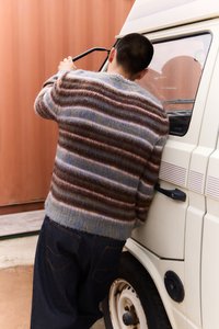 Pletený svetr s horizontálními pruhy v hnědé, krémové a modré barvě; volný střih; kombinován s tmavými džínami. V pozadí bílý vůz.