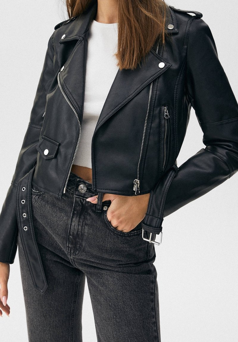 Veste de motard en cuir noir au design court, avec des accessoires argentés, des poches zippées et un détail ceinturé. Portée sur un haut blanc, assortie à un jean foncé.