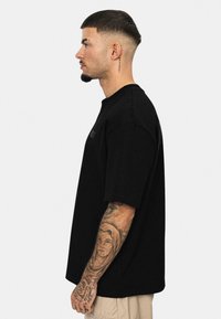 Sort oversized bomulds t-shirt med korte ærmer og rund hals; broderet logo på brystet; modellen har tatoveringer og kort hår.