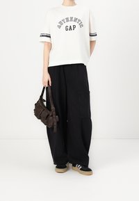 Bílá tričko s potiskem "AUTHENTIC GAP", černé široké kalhoty a hnědá taška přes rameno. Černé tenisky s béžovými podrážkami a třemi pruhy.
