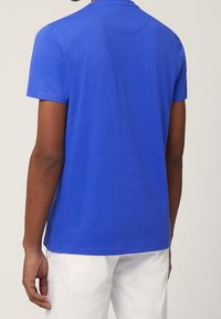 Blå t-shirt med kort ärm tillverkad av slät tyg. Rund halsringning med en rund sömdetalj på baksidan. Bärs med vita byxor.