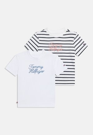 To børneskjorter med korte ærmer med teksten "Tommy Hilfiger"; en ensfarvet hvid med blå skrift, en hvid med sorte striber og rød skrift.
