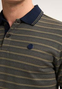 Pruhovaná polo košile v námořnické modré a olivové zelené. Obsahuje jednobarevný námořnický límeček se dvěma knoflíky a malý značkový detail na hrudi.