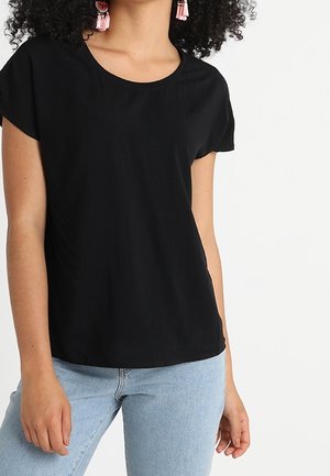Femme portant un t-shirt noir à manches courtes, un jean bleu clair et des boucles d'oreilles à pampilles roses sur un fond blanc.