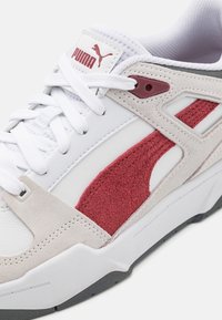 Bílé sportovní tenisky s červenými semišovými akcenty, hladkou kůží a texturovaným síťovaným jazykem. Šněrování se značením Puma na jazyku.