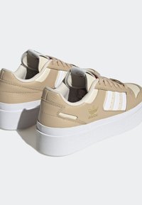 Beige ja valged platvormiga Adidas spordijalatsid valgete taldadega, ikoonilised kolm triipu ja kuldne Adidas logo küljel.