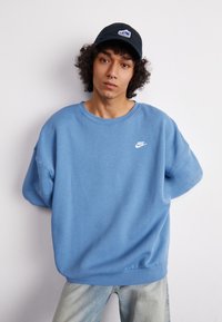 Fénykék, túlgenerált pulóver kerek kivágással, fehér Nike logóval a mellkason. Fekete sapkával párosítva.