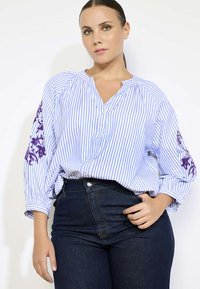 Chemisier à rayures bleues et blanches avec une broderie florale violette sur les manches. Présente une coupe décontractée et un décolleté en V.