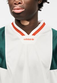 Αθλητική φανέλα Adidas με λευκό σώμα, πράσινα μανίκια από δίχτυ και κόκκινο γιακά με διακοσμητική λεπτομέρεια· το λογότυπο εμφανίζεται στο στήθος.