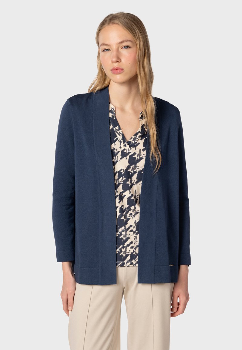 Marinblå cardigan i texturerat tyg, med avslappnad passform, öppen framsida och långa ärmar, lager-på-lager över en mönstrad blus.