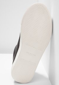 Baskets noires avec une semelle en caoutchouc texturée, présentant un motif en zigzag. Design minimaliste avec un bord blanc contrastant. Nom de la marque embossé sur la semelle.