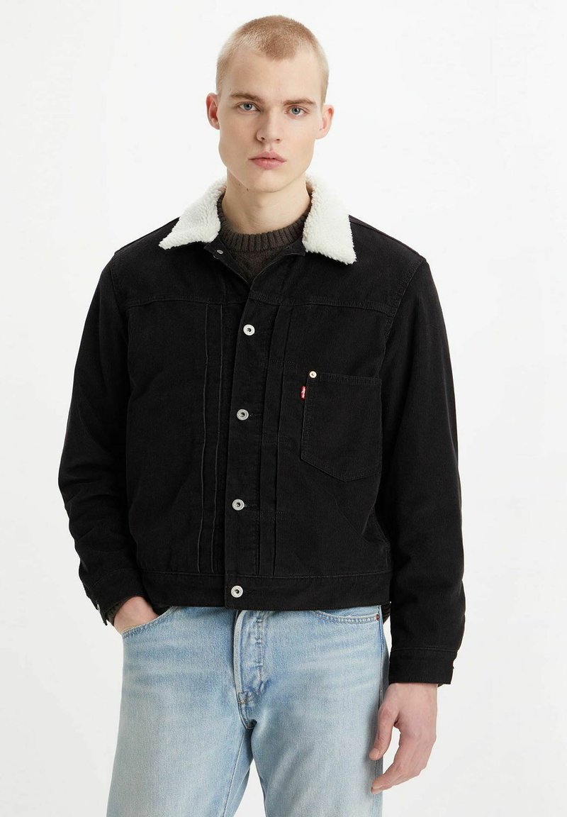 Black corduroy jacket with a white fleece collar, featuring a button fastening, front pocket, and a relaxed fit. Worn with light blue jeans.