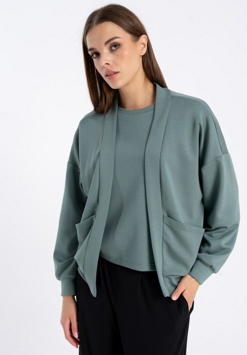 Jasnozielona, oversize'owa kurtka z polarowego materiału, z długimi rękawami, otwartym przodem i dwiema przednimi kieszeniami, noszona na dopasowanej koszuli i czarnych spodniach.