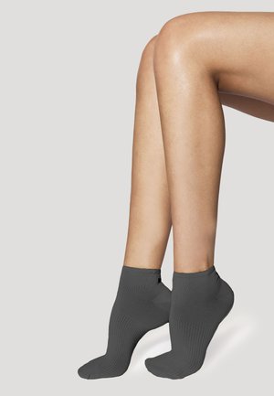 Jambes levées sur la pointe des pieds, portant des chaussettes côtelées grises chevillées sur un fond clair et neutre.