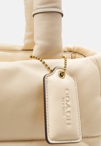Kremowa skórzana torebka o gładkiej fakturze, zaokrąglonych uchwytach oraz złotym łańcuszkiem z wytłoczonym logo marki.