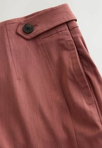 Close-up van roestkleurige stoffen broek met een met knopen bevestigd zijtabblad en zichtbare zakdetail.