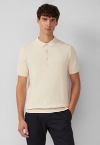 Polo w kolorze kremowym z krótkimi rękawami, z teksturowanym, skośnym wzorem w paski, trzema guzikami z przodu oraz ściągaczem przy kołnierzy.
