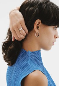 La joyería de plata incluye un anillo liso y un arete de aro delgado. El modelo lleva una blusa azul con plisados y un cuello alto.