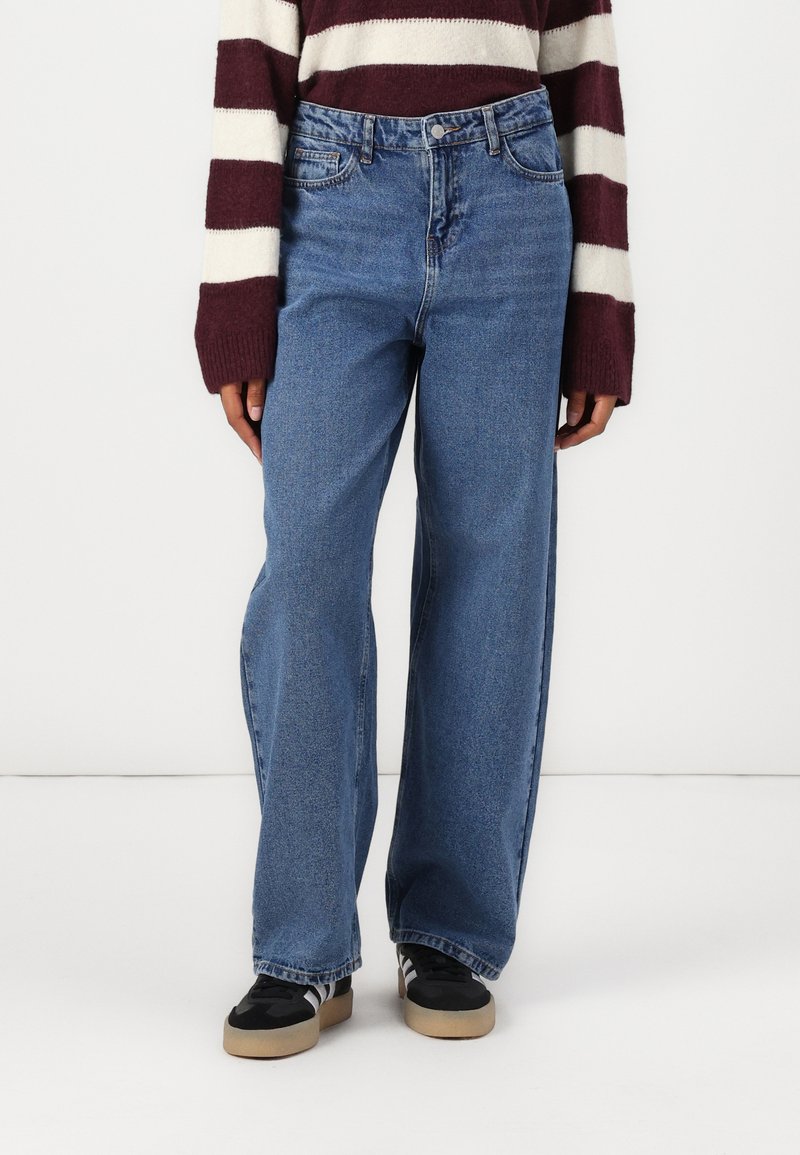 Weite blaue Jeans mit hoher Taille, ausgestattet mit Vordertaschen und einem klassischen Knopfverschluss. Kombiniert mit schwarz-weißen Sneakers.
