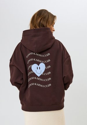 Hoodie oversize marrone con un grande cuore blu e la scritta "GOOD KARMA CLUB" in bianco, esposta in un motivo spirale sul retro.