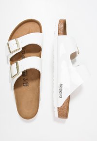 Weiße Birkenstock-Sandalen mit zwei verstellbaren Schnallen, einem Korkfußbett und einer strukturierten Sohle. Glatte, glänzende Lackoberfläche mit leichtem Glanz.