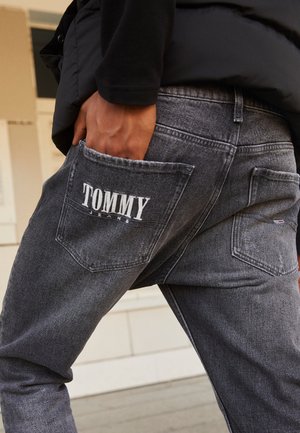 Personne portant un jean noir avec la main dans la poche arrière, logo « TOMMY JEANS » visible sur la poche arrière droite dans un cadre décontracté.