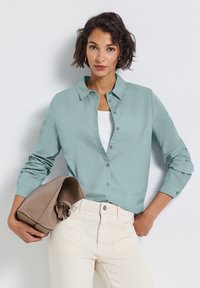Donna che indossa una camicia azzurra chiara abbottonata sopra una maglietta bianca, pantaloni avorio, tiene sotto il braccio una borsa piegata color taupe su uno sfondo bianco.