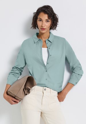 Femme portant une chemise bleu clair boutonnée sur un top blanc, un pantalon ivoire, tenant un sac taupe plié sous son bras contre un fond blanc.