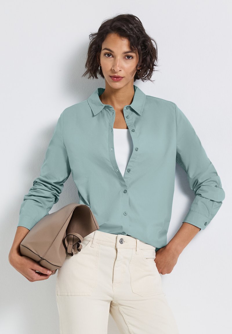 Vrouw in een lichtblauwe blouse over een witte top, met off-white broek, die een opgevouwen taupe tas onder haar arm houdt tegen een witte achtergrond.