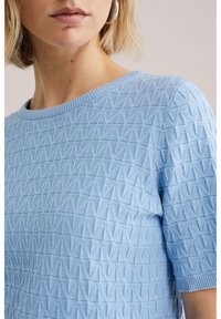 Vaaleansininen neulottu paita, jossa kohokuvioitu timanttimainen kuviointi, pyöreä pääntie ja lyhyet hihat, yksinkertaisella resorireunuksella.