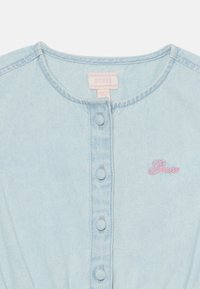 Haut en denim bleu clair avec un col rond, fermeture à boutons sur le devant, logo "Guess" brodé en rose et texture de tissu douce.