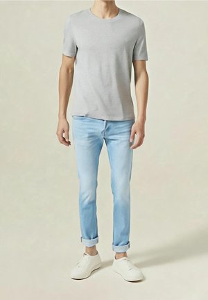 Mężczyzna ubrany w jasnoszarą koszulkę z krótkim rękawem, jasnoniebieskie podwinięte jeansy i białe trampki, stojący na neutralnym tle.