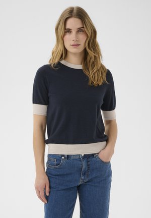 Jeune femme aux cheveux blonds porte un pull à manches courtes bleu marine avec une bordure beige et un jean bleu, debout avec une main dans la poche.