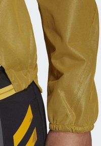 Gele sportjas met mesh mouw en elastische manchetten, gecombineerd met zwarte legging met gele en grijze diagonale strepen.