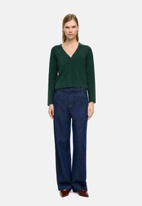 Zielony sweter zapinany na guziki z dekoltem w kształcie litery V. Połączony z luźnymi, ciemnoniebieskimi dżinsami i brązowymi spiczastymi butami.