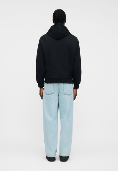 Άνδρας που στέκεται με την πλάτη στραμμένη, φοράει μια μαύρη φούτερ με κουκούλα και ανοιχτό μπλε παντελόνι φαρδιάς γραμμής με μαύρα παπούτσια, σε απλό φόντο.