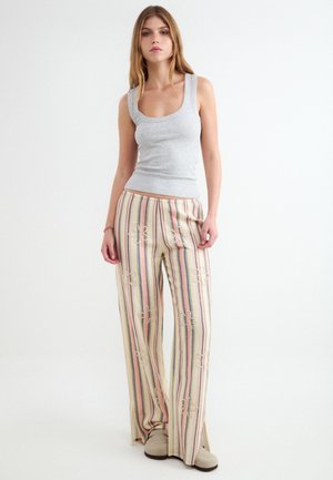 Giovane donna in piedi che indossa una canotta grigia e pantaloni larghi a righe multicolori con scarpe beige su sfondo bianco.