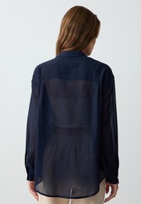 Woman with brown hair wearing a sheer navy blue long-sleeve shirt over a black top, facing away against a plain white background.