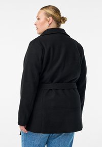 Schwarzer Mantel mit Gürtel in der Taille, eingeschnittenem Kragen und taillierter Passform. Der Stoff wirkt glatt und weist ein klares Design ohne Verzierungen auf.